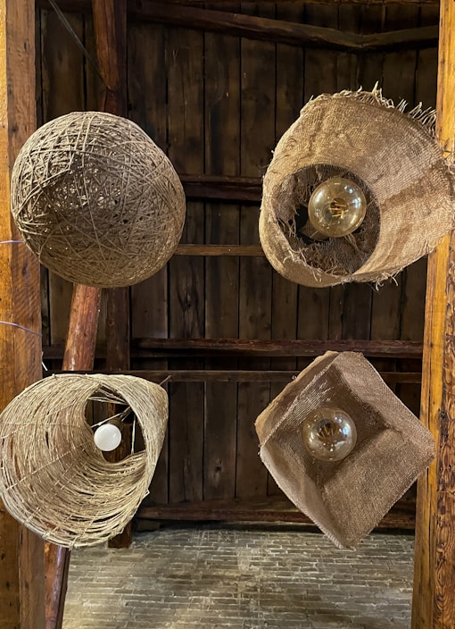 Multiple rustic light fixtures hang from a wooden ceiling, each made from natural materials like twine and burlap. The fixtures feature exposed bulbs, adding to the organic and handmade aesthetic, set against a backdrop of dark wooden planks.