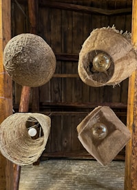 Multiple rustic light fixtures hang from a wooden ceiling, each made from natural materials like twine and burlap. The fixtures feature exposed bulbs, adding to the organic and handmade aesthetic, set against a backdrop of dark wooden planks.