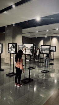 A person, wearing a hijab and a pink shirt, is pointing at a picture in an art gallery. Several black and white photographs are displayed on stands in a well-lit room with reflective flooring. Other individuals are seen in the background, observing the photographs.