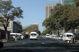 A busy urban street scene with several white minivans parked alongside the road. Tall buildings are visible in the background amid lush green trees lining the street. A few pedestrians are walking on the sidewalks, and traffic lights can be seen overhead.