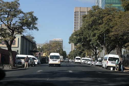 A busy urban street scene with several white minivans parked alongside the road. Tall buildings are visible in the background amid lush green trees lining the street. A few pedestrians are walking on the sidewalks, and traffic lights can be seen overhead.