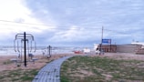 Outdoor gym equipment is set up on a grassy area near a beach. A walkway made of pavers leads towards the ocean. The sky appears overcast, and the waves in the background indicate a windy atmosphere. A sign with the name 'Nautilus' is visible near a building.