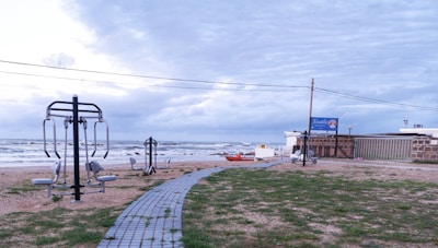 Outdoor gym equipment is set up on a grassy area near a beach. A walkway made of pavers leads towards the ocean. The sky appears overcast, and the waves in the background indicate a windy atmosphere. A sign with the name 'Nautilus' is visible near a building.