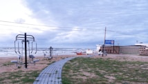 Outdoor gym equipment is set up on a grassy area near a beach. A walkway made of pavers leads towards the ocean. The sky appears overcast, and the waves in the background indicate a windy atmosphere. A sign with the name 'Nautilus' is visible near a building.
