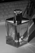 A glass perfume bottle with a metallic cap, featuring the label 'H24'. The background includes a keyboard, indicating a desk setting. The lighting creates reflections and highlights on the bottle.