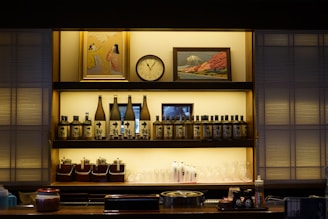 A well-organized shelf featuring bottles of sake, condiment containers, glasses, and some decor items such as a clock and framed artwork. The shelf and its contents are illuminated with soft lighting, creating a cozy atmosphere. The artwork above includes a painting of people and another of a landscape, possibly a mountain.