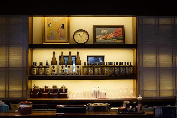 A well-organized shelf featuring bottles of sake, condiment containers, glasses, and some decor items such as a clock and framed artwork. The shelf and its contents are illuminated with soft lighting, creating a cozy atmosphere. The artwork above includes a painting of people and another of a landscape, possibly a mountain.