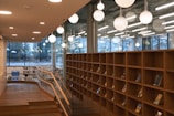A modern library or bookstore interior featuring tall wooden bookshelves partially filled with books. Large, spherical pendant lights hang from the ceiling, providing warm illumination. Glass walls with large windows allow natural light to enter and offer views of the outdoors. A wooden railing borders a stepped path leading through the space.