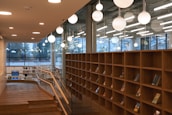 A modern library or bookstore interior featuring tall wooden bookshelves partially filled with books. Large, spherical pendant lights hang from the ceiling, providing warm illumination. Glass walls with large windows allow natural light to enter and offer views of the outdoors. A wooden railing borders a stepped path leading through the space.