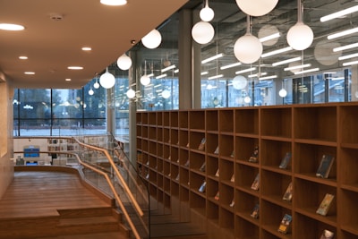 A modern library or bookstore interior featuring tall wooden bookshelves partially filled with books. Large, spherical pendant lights hang from the ceiling, providing warm illumination. Glass walls with large windows allow natural light to enter and offer views of the outdoors. A wooden railing borders a stepped path leading through the space.