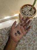 A hand with black ink drawings or symbols on the palm is resting on a patterned fabric. Nearby, there is a potted plant with white pebbles on the surface. The pot is placed near a wall with subtle lighting.