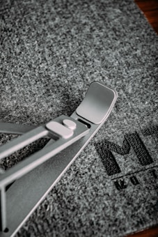A metallic stand with a triangular support structure is placed on a textured gray mat, which has partial text printed on it. The stand features a modern design with sleek lines and a hinge mechanism.