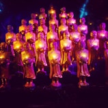 A group of small statues depicting monks holding illuminated objects, arranged in rows. The statues are bathed in vibrant purple and gold lighting, creating a mystical atmosphere.