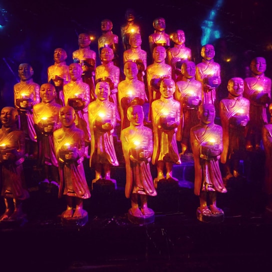 A group of small statues depicting monks holding illuminated objects, arranged in rows. The statues are bathed in vibrant purple and gold lighting, creating a mystical atmosphere.