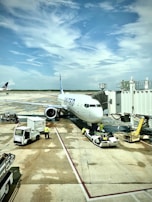 A commercial airplane is parked at an airport gate with a jet bridge connected to it. Ground crew members are visible preparing the aircraft, with various service vehicles such as baggage carts and tow tractors nearby. The sky is partly cloudy, providing a backdrop with expanses of blue and white.