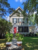A charming two-story house with a gable roof surrounded by lush greenery. The exterior features light-colored siding with black shutters. A bright red front door stands out against the darker shades, and two large pots of vibrant red flowers line the walkway leading up to the entrance. Mature trees and well-maintained shrubs frame the setting, creating a peaceful and inviting atmosphere.