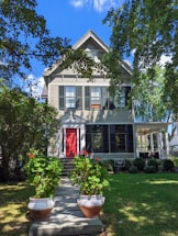 A charming two-story house with a gable roof surrounded by lush greenery. The exterior features light-colored siding with black shutters. A bright red front door stands out against the darker shades, and two large pots of vibrant red flowers line the walkway leading up to the entrance. Mature trees and well-maintained shrubs frame the setting, creating a peaceful and inviting atmosphere.