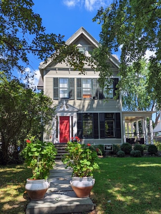A charming two-story house with a gable roof surrounded by lush greenery. The exterior features light-colored siding with black shutters. A bright red front door stands out against the darker shades, and two large pots of vibrant red flowers line the walkway leading up to the entrance. Mature trees and well-maintained shrubs frame the setting, creating a peaceful and inviting atmosphere.