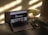 A laptop displaying a webpage sits on a dark table with an open internet browser. Beside the laptop is a transparent vase filled with white flowers. A smartphone lies next to the vase, reflecting the shadows and light on the table.