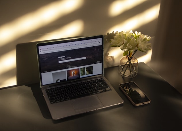 A laptop displaying a webpage sits on a dark table with an open internet browser. Beside the laptop is a transparent vase filled with white flowers. A smartphone lies next to the vase, reflecting the shadows and light on the table.