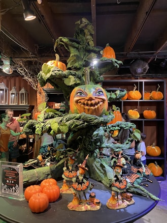 A large, whimsical Halloween display features a central tree structure adorned with intricately carved pumpkins and lush green leaves. The focal point is a large pumpkin with a mischievous face at the center. Smaller pumpkin figurines and decorations, including witches, are scattered around. The area is warmly lit with soft yellow lighting, creating a playful and festive atmosphere.