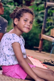 A young child with curly hair, wearing a patterned top and pink shorts, sitting outdoors on a woven mat. The background features lush greenery, suggesting a natural setting. The child has a calm and content expression.