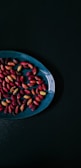 A collection of colorful, oval-shaped seeds are scattered on a dark blue, round plate. The seeds exhibit a variety of rich colors, predominantly reds and purples with some hints of yellow. The plate is placed against a dark, almost black background, creating a stark contrast that emphasizes the vibrant colors of the seeds.