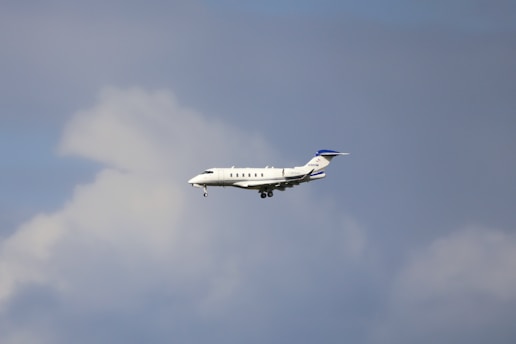 A mid-sized private jet flying through a cloudy sky, showing its landing gear extended.