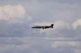 A sleek private jet is flying through a cloudy sky. The aircraft is dark in color and its landing gear is extended. The clouds vary from white to gray, creating a dramatic backdrop.