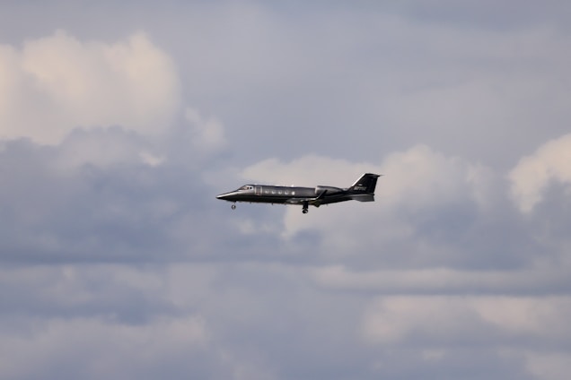 A sleek private jet is flying through a cloudy sky. The aircraft is dark in color and its landing gear is extended. The clouds vary from white to gray, creating a dramatic backdrop.