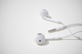 A pair of white wired earphones with a 3.5mm audio jack lying on a light surface. The earphones are designed for in-ear use and have a sleek, minimalist appearance.