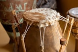 A traditional drum with a wooden base and an animal skin head. The drum is secured with strings tied to wooden pegs. In the background, there are other drums and pottery with intricate designs.