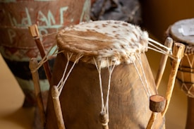 A traditional drum with a wooden base and an animal skin head. The drum is secured with strings tied to wooden pegs. In the background, there are other drums and pottery with intricate designs.