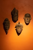 Four traditional wooden masks are mounted on an orange wall. The masks are dark in color with distinct carved features, such as prominent noses, closed eyes, and tribal markings.