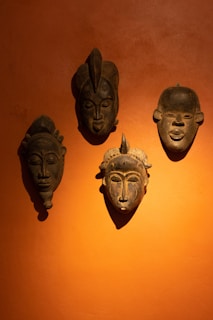 Four traditional wooden masks are mounted on an orange wall. The masks are dark in color with distinct carved features, such as prominent noses, closed eyes, and tribal markings.