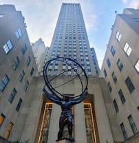 A large, imposing skyscraper with numerous windows dominates the background, framed by additional tall buildings on either side. In the foreground, a massive statue of Atlas is depicted holding up a large armillary sphere, symbolizing strength and endurance.