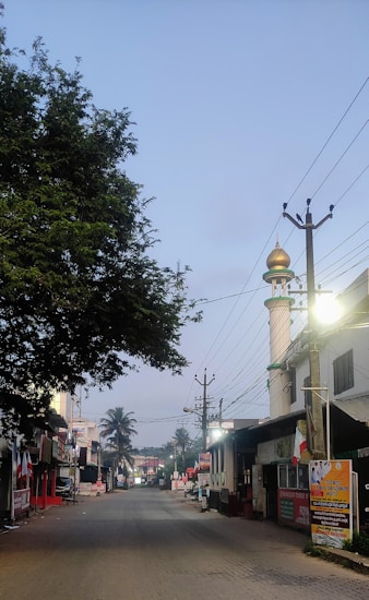 A quiet street scene with shops and buildings lining the road. A mosque with a recognizable dome and minaret is visible on the right side. Trees provide shade and power lines stretch across the sky. A few streetlights are illuminated, casting a warm glow on the surroundings.