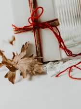 A white gift box intricately tied with red yarn and string, complemented by a delicate lace fabric. The backdrop includes autumn leaves scattered gracefully around the box.
