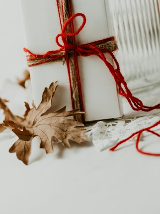 A white gift box intricately tied with red yarn and string, complemented by a delicate lace fabric. The backdrop includes autumn leaves scattered gracefully around the box.