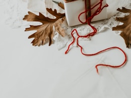 An arrangement featuring a white gift box wrapped in twine and red yarn, flanked by two brown oak leaves on a lace background.