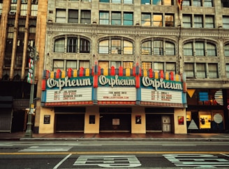 A historic theater with a grand marquee displaying the name 'Orpheum', bordered by intricate architectural details and large windows. The marquee lists upcoming events or performers, surrounded by vintage-style design elements.