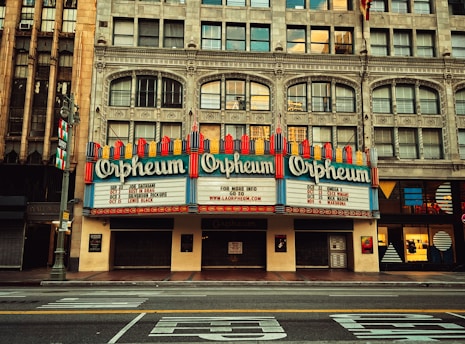 A historic theater with a grand marquee displaying the name 'Orpheum', bordered by intricate architectural details and large windows. The marquee lists upcoming events or performers, surrounded by vintage-style design elements.