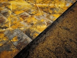 A close-up view of a textured surface showing wet, square tiles with a mix of yellow, orange, brown, and gray patterns. Adjacent to the tiles is a section of loose granular material, possibly sand or soil, partially covering the base of the tiles.