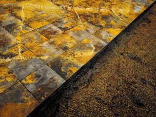 A close-up view of a textured surface showing wet, square tiles with a mix of yellow, orange, brown, and gray patterns. Adjacent to the tiles is a section of loose granular material, possibly sand or soil, partially covering the base of the tiles.