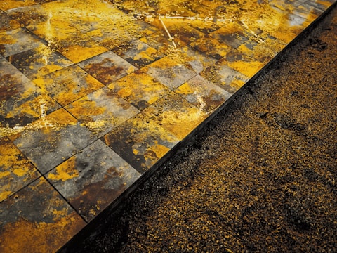 A close-up view of a textured surface showing wet, square tiles with a mix of yellow, orange, brown, and gray patterns. Adjacent to the tiles is a section of loose granular material, possibly sand or soil, partially covering the base of the tiles.