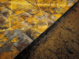 A close-up view of a textured surface showing wet, square tiles with a mix of yellow, orange, brown, and gray patterns. Adjacent to the tiles is a section of loose granular material, possibly sand or soil, partially covering the base of the tiles.