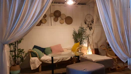 A cozy indoor setting featuring a small seating area with a couch adorned with cushions of various colors. A glass-top coffee table sits in front of the couch, accompanied by a soft footstool. The walls display decorative woven baskets and a dreamcatcher. Soft lighting and plants enhance the tranquil ambiance, while large, flowing curtains create an intimate atmosphere.