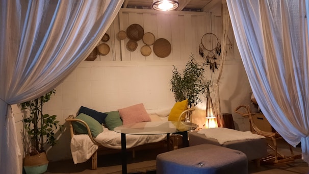 A cozy indoor setting featuring a small seating area with a couch adorned with cushions of various colors. A glass-top coffee table sits in front of the couch, accompanied by a soft footstool. The walls display decorative woven baskets and a dreamcatcher. Soft lighting and plants enhance the tranquil ambiance, while large, flowing curtains create an intimate atmosphere.