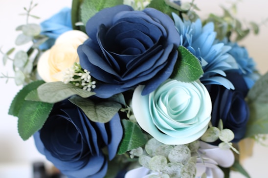 A close-up view of a bouquet composed of artificial flowers, including dark blue, light blue, and pale yellow roses along with green leaves and small white floral accents.