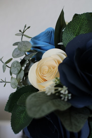 A close-up of artificial flowers featuring intricately crafted paper roses in blue and cream colors, accompanied by green leaves and delicate white buds. The arrangement combines realistic textures with a soft and elegant presentation.